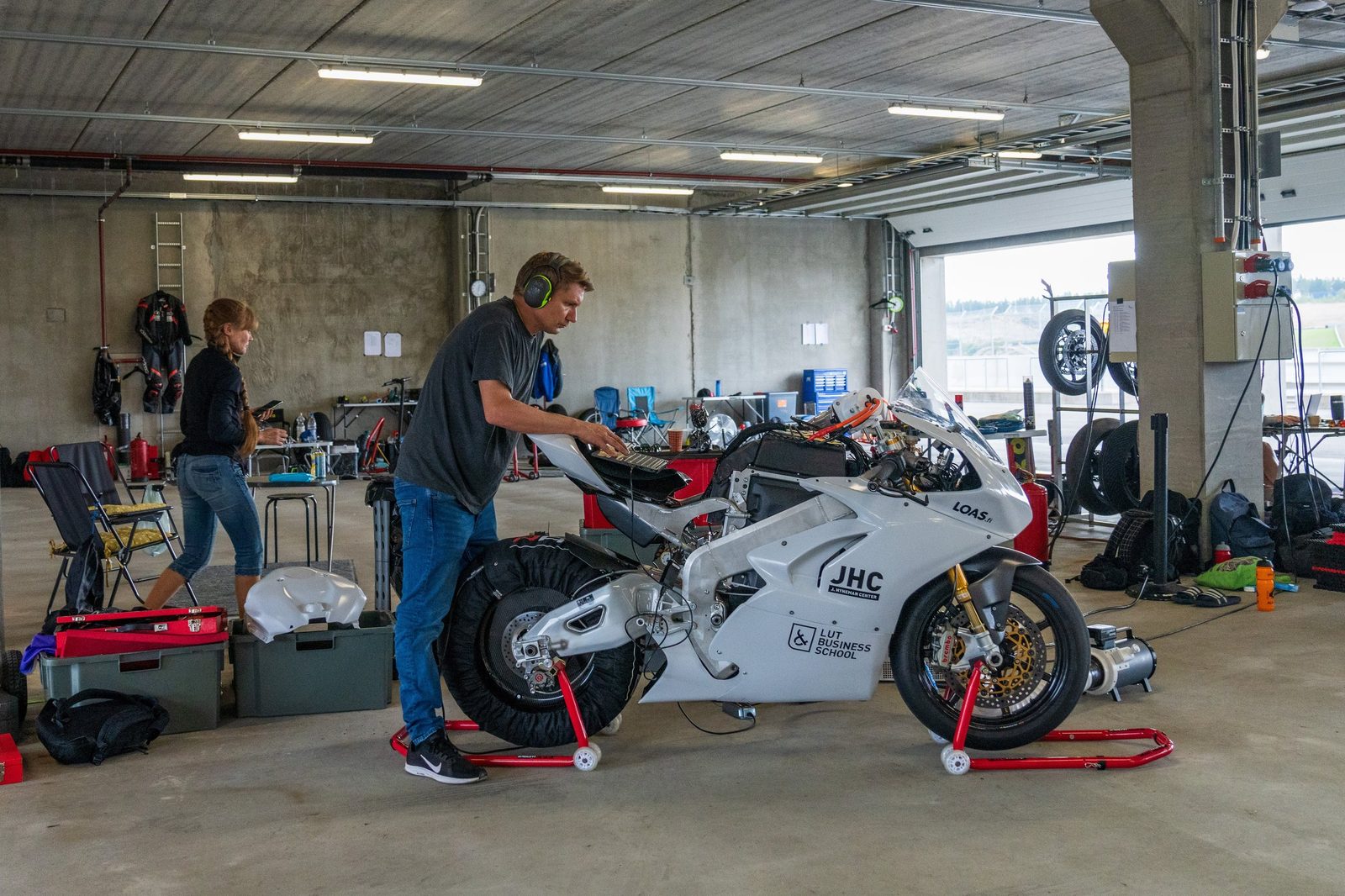 Mk3 on paddock stands in the KymiRing garage with a leaf blower on the floor aimed into the bodywork, and Marko Kasurinen at a laptop on the seat unit.