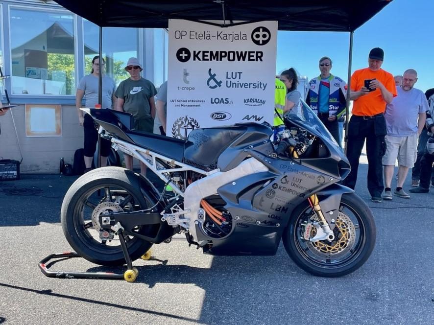 Mk1 on a paddock stand under a JHC awning at Lappeenranta airfield, sponsor board behind it, a small crowd around.