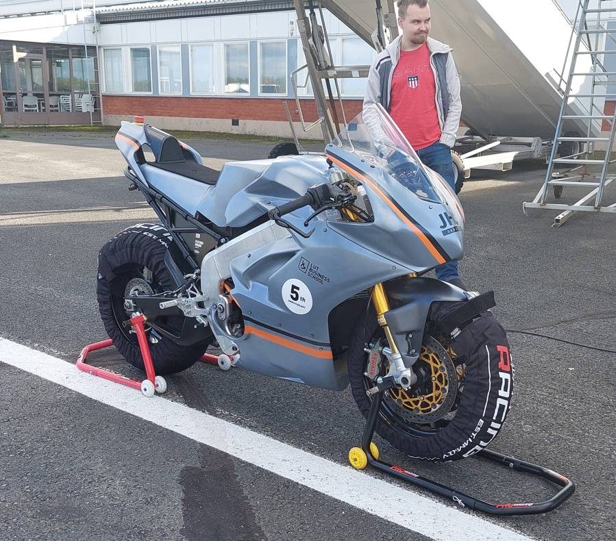 Mk2 on a paddock stand at the airfield with tyre warmers fitted, and test rider Jere Honkanen standing behind it.