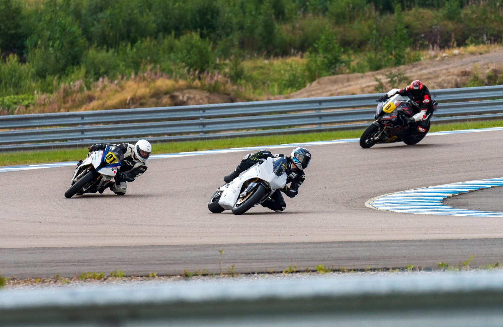 Mk3 leaned over mid-corner at KymiRing, with a combustion motorcycle ahead of it and another behind on the same stretch of track.